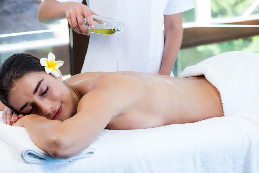 Une femme se détend lors d'un massage thaïlandais à l'huile dans un spa, les yeux fermés et un sourire serein. Une praticienne en uniforme blanc verse de l'huile sur son dos, avec une fleur de frangipanier ornant ses cheveux, créant une atmosphère de bien-être et de relaxation.